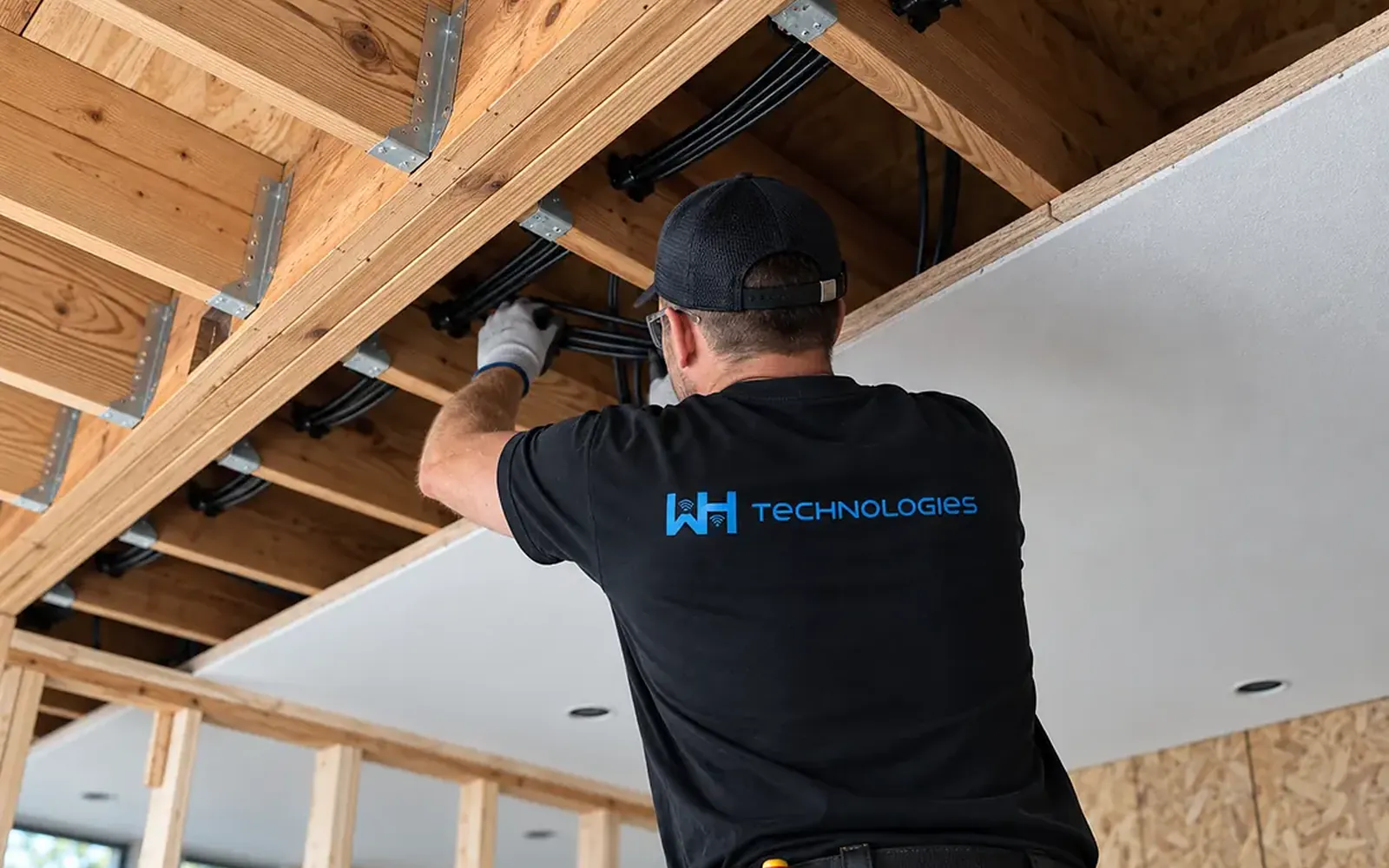 WH Technologies technician on a ladder fixing a round white access point to a stained wood ceiling, with the cable already run to the mounting point