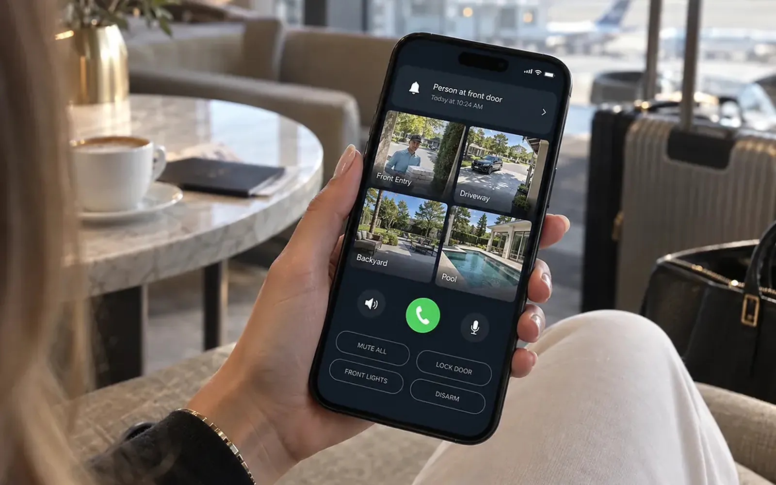 Hand holding a smartphone at a cafe table, the screen showing a short list of camera alerts with thumbnails of a front door and a driveway