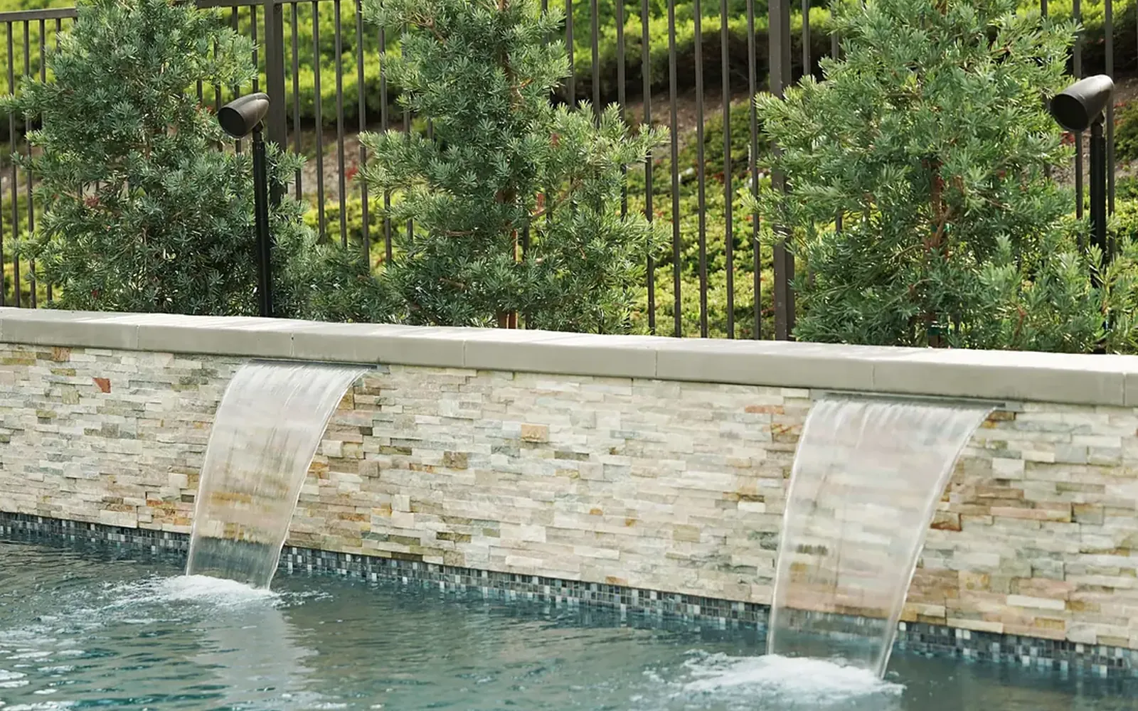 Pool water feature built from natural stone with a rock-style landscape speaker set among the boulders, nearly indistinguishable from them