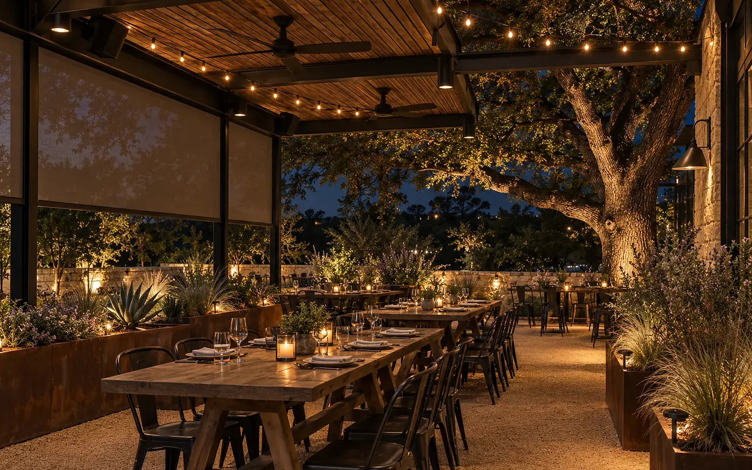 Covered restaurant patio at night under an uplit tree: steel pergola with string lights and fans, small outdoor speakers under the beams, a motorized screen lowered halfway, corten planters with agaves and grasses