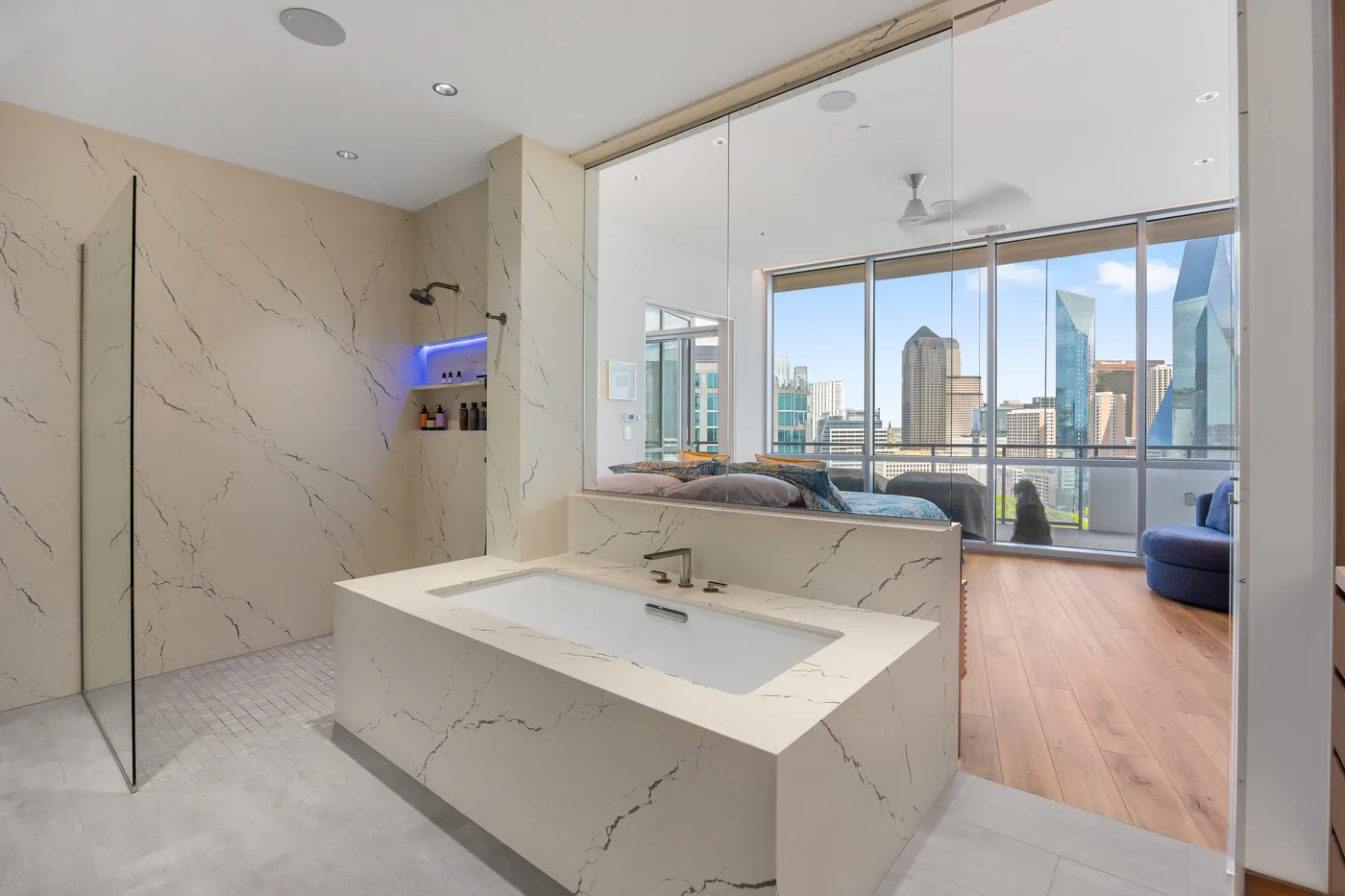 Freestanding tub by floor-to-ceiling glass with the skyline, a lit mirror and a lit vanity