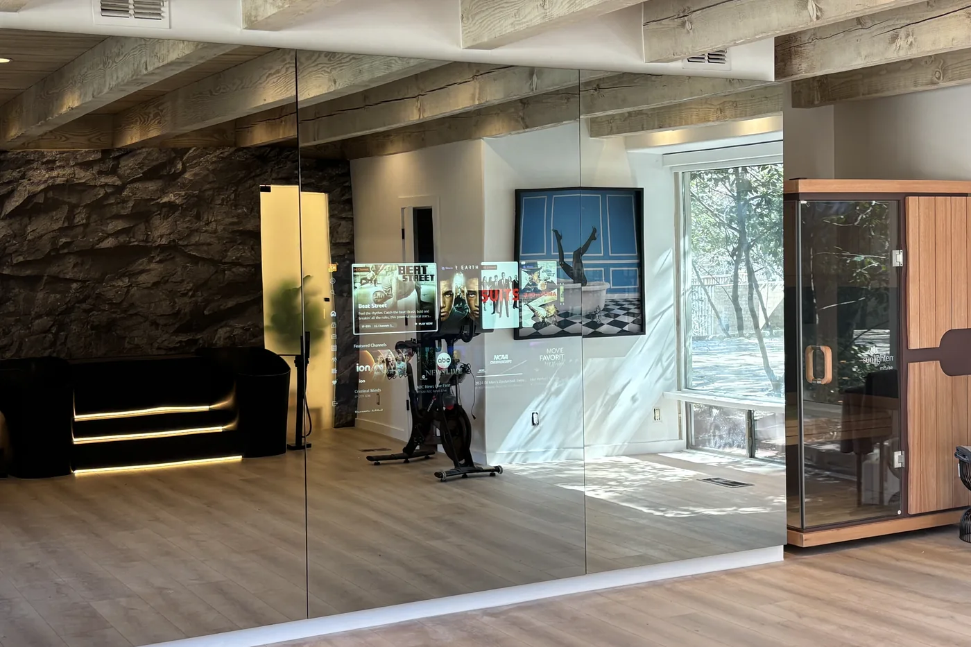 Home gym with a mirror TV showing a workout, a stone accent wall and windows to the trees