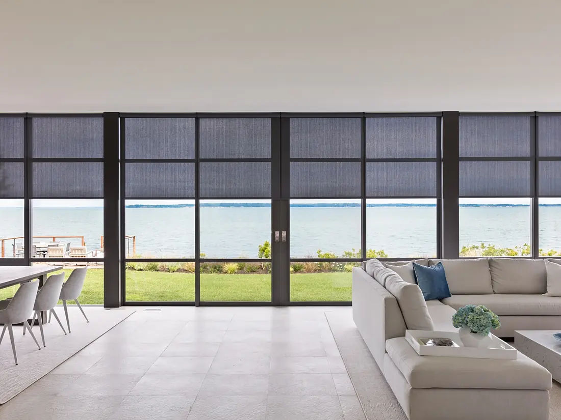 Open-plan living room with a wall of glass and dark solar shades lowered against the sun, with the water view still visible